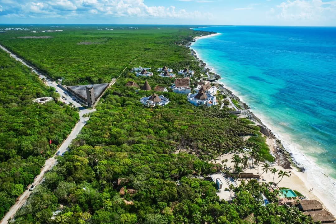 Aerial view showing a lush green landscape with a beach and turquoise ocean in the background, along with coastal buildings and roads.$#$ CAPTION
