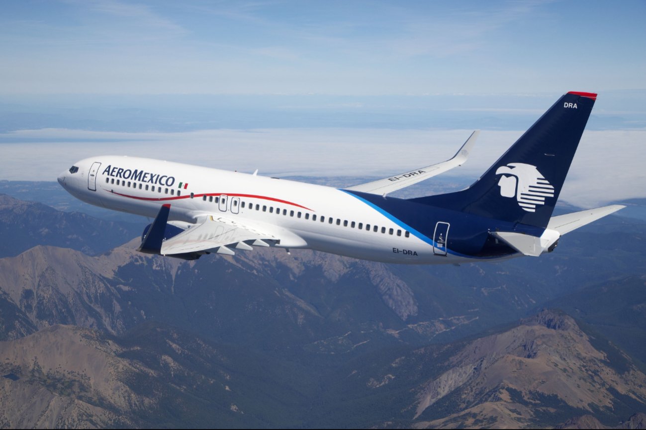 A sleek Aeroméxico airplane flying over a mountainous landscape with blue skies in the background.$#$ CAPTION