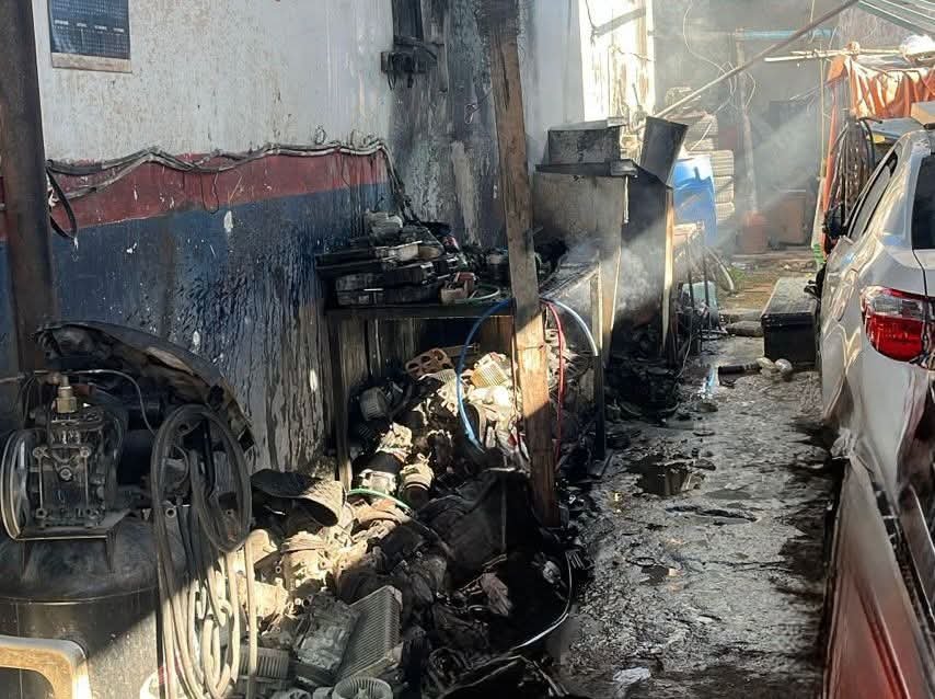 A narrow passage in an auto repair shop showing burnt debris, damaged machinery, and smoke lingering in the air after a fire incident.$# CAPTION
