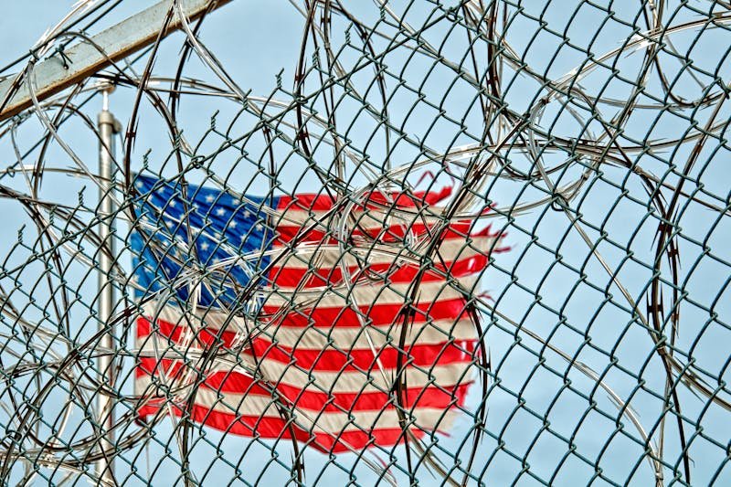 An American flag waving in the background, partially obscured by a barbed wire fence