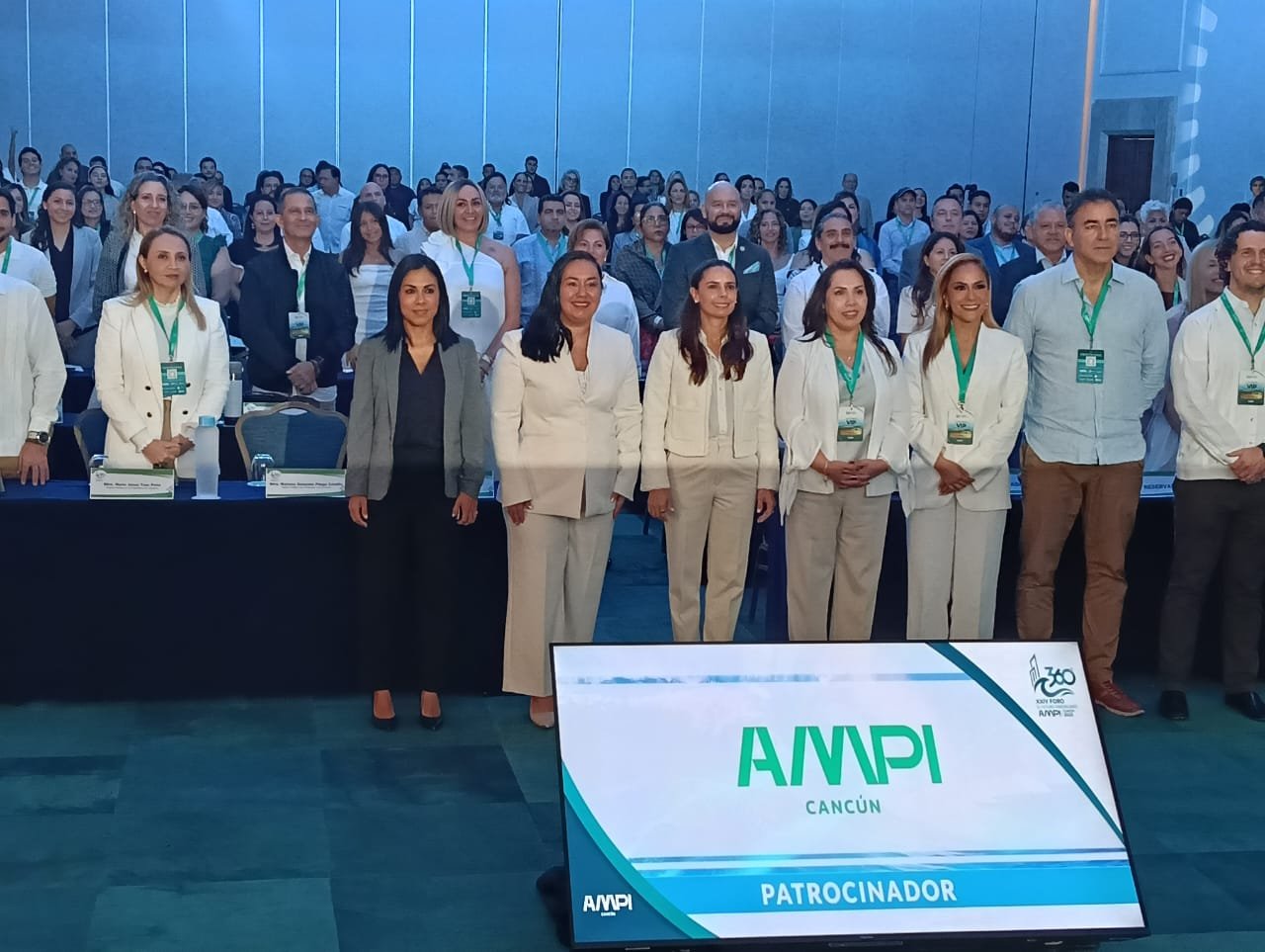 A group of attendees stands in front of a panel at the AMPI conference in Cancun with a large display screen showing the AMPI logo and the word Patrocinador