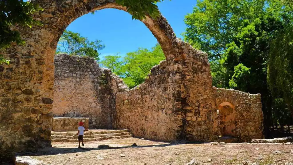 a person standing in front of stone ruins with an arch under a bright blue sky and trees in the background