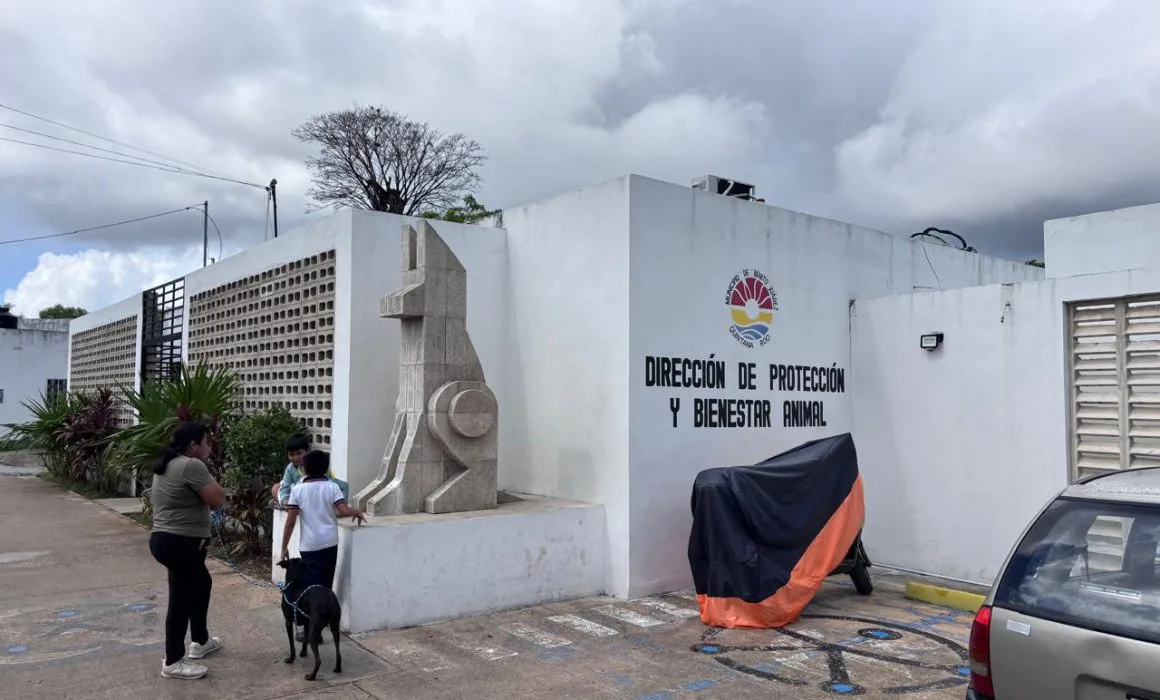 A woman and a child stand outside an animal protection center with a statue of a dog and a parked car. A dog is with them.