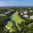 Aerial view of a golf course with a winding waterway and green fairways surrounded by residential buildings and trees under a clear blue sky.$#$ CAPTION