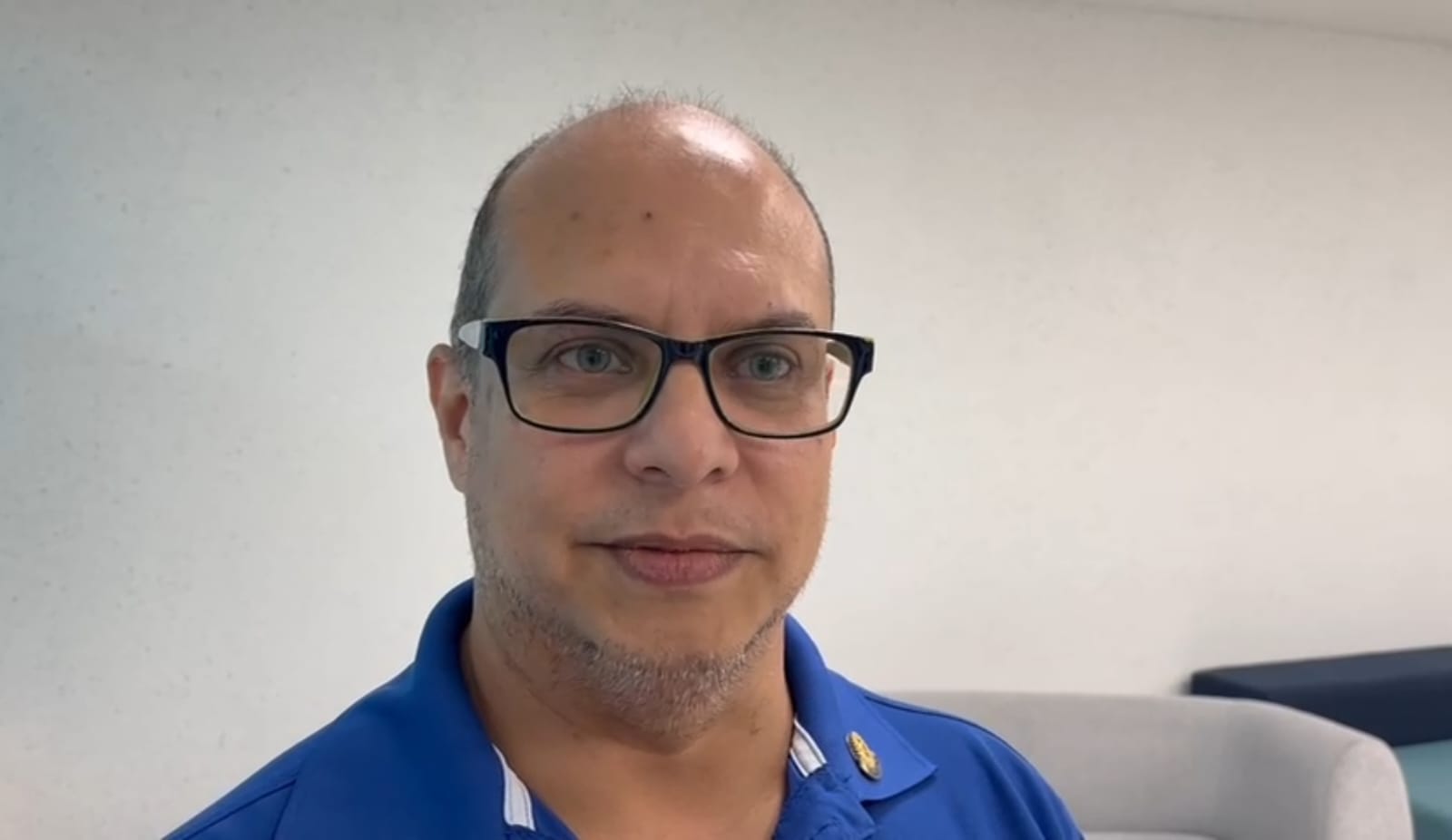 A man with glasses and a blue shirt looking thoughtfully at the camera in a well-lit room with a light-colored background.$