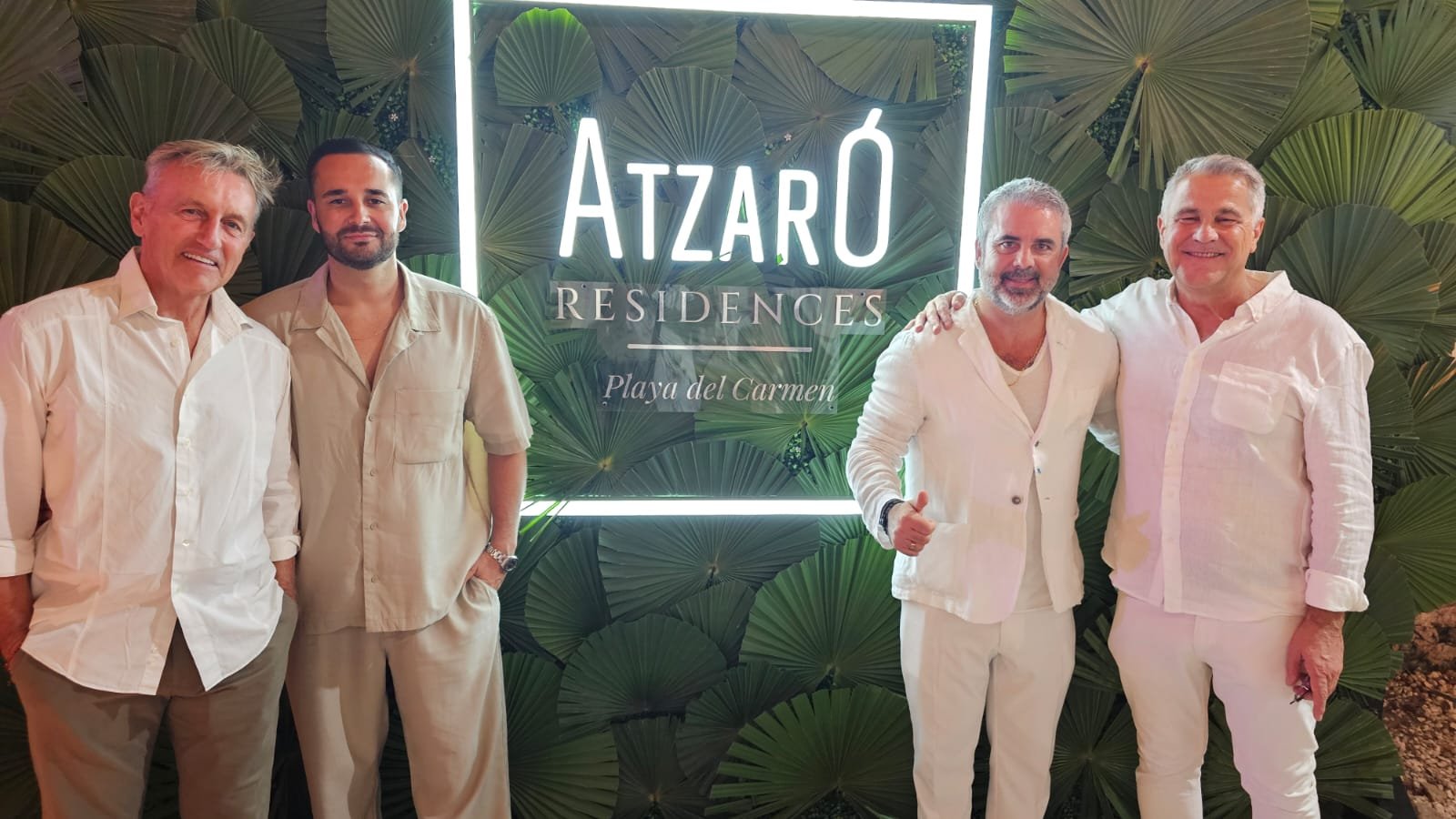 A group of four men dressed in light-colored attire posing for a photo in front of a tropical backdrop with the Atzaró Residences logo.