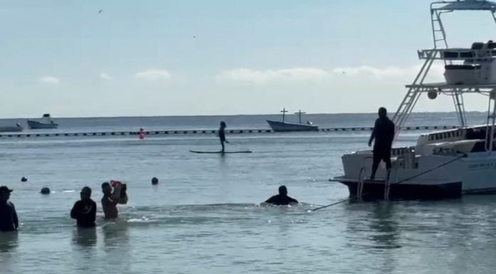 A scenic view of a beach with people swimming, paddleboarding, and a boat nearby under a clear sky.