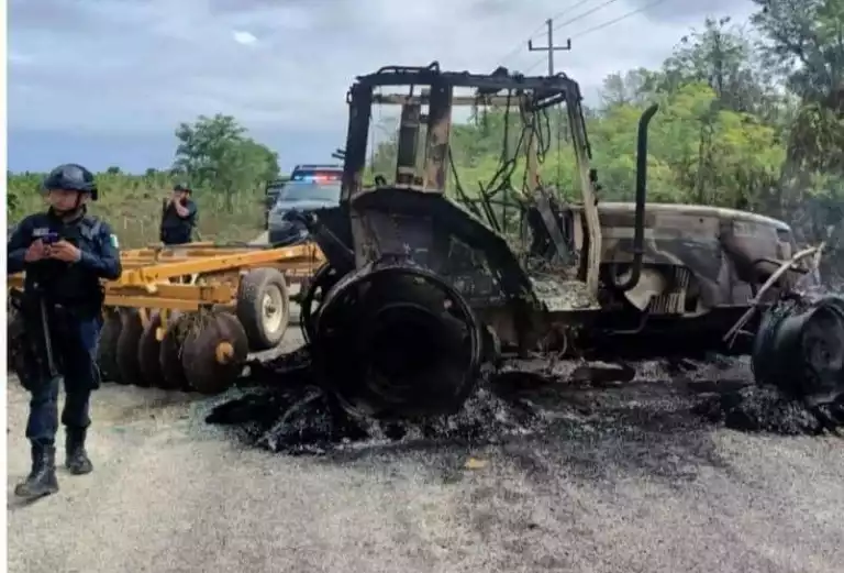 A burnt tractor and farming equipment on a roadside, with police officers present in the background.$# CAPTION