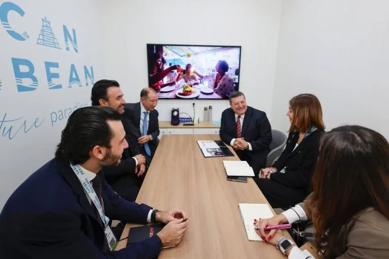 a group of professionals sitting around a conference table engaged in discussion with a television screen showing a dining scene in the background