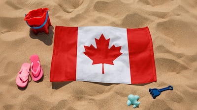 A Canadian flag surrounded by beach toys on sand, including a bucket, pink flip-flops, and some plastic items.$
