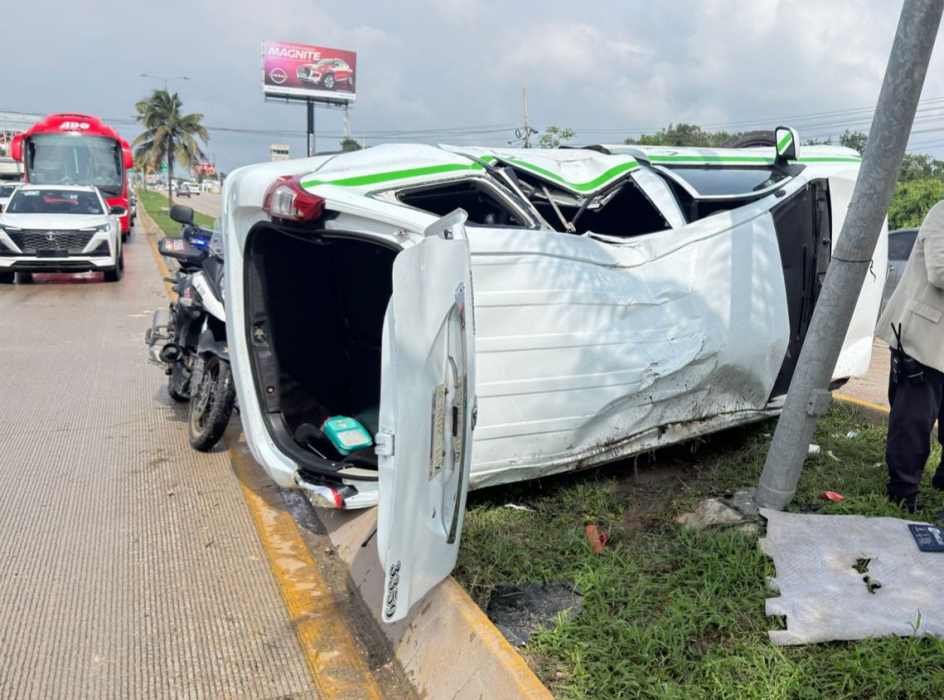 An overturned white vehicle on the roadside, with its door open and surrounding traffic visible in the background. A motorcycle is parked nearby, and vegetation is present on the roadside. A billboard is seen in the distance.
