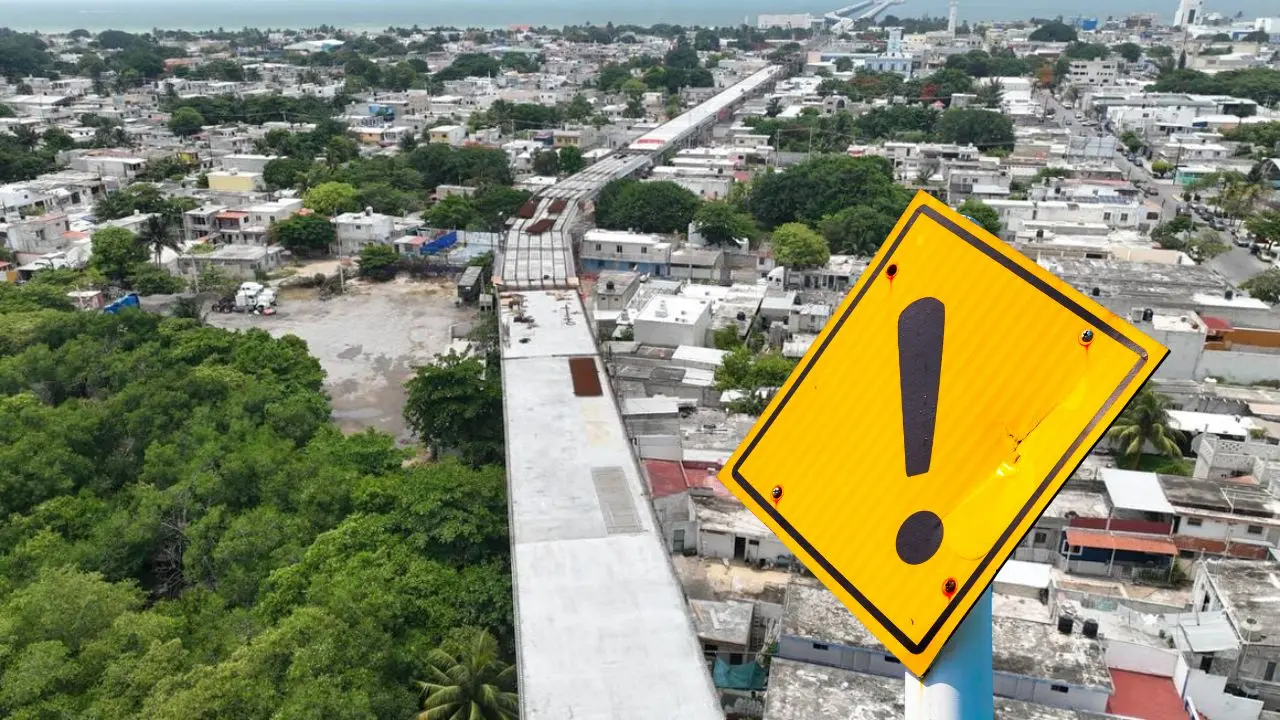 A bright yellow caution sign with an exclamation mark stands in the foreground, overlooking a densely populated urban area with residential buildings and greenery in the background. A train track runs through the scene.