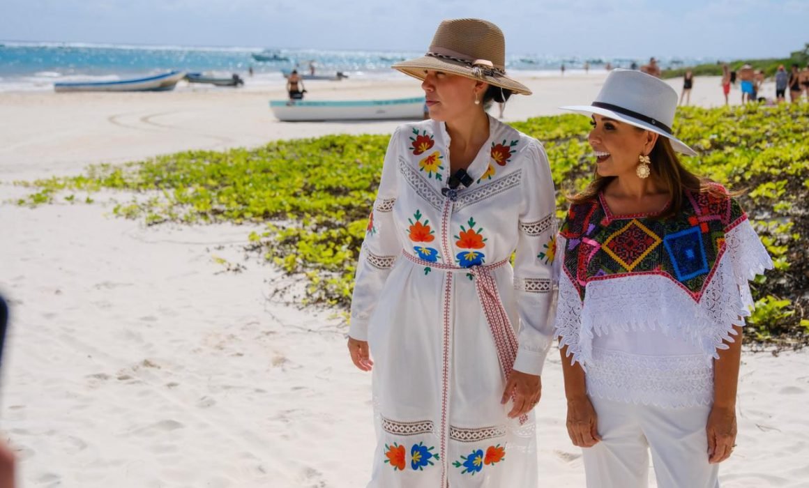 Two women in traditional embroidered clothing stand on a beach with a lush green area in the foreground and the ocean in the background.$# CAPTION