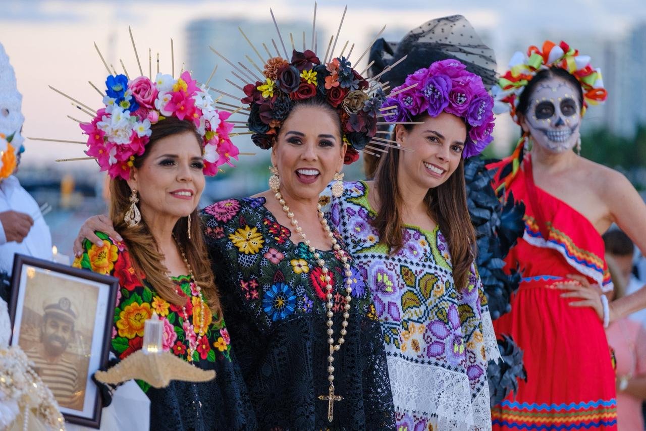 Women dressed in colorful traditional attire with floral crowns, participating in a cultural celebration, while holding a framed photograph.$# CAPTION