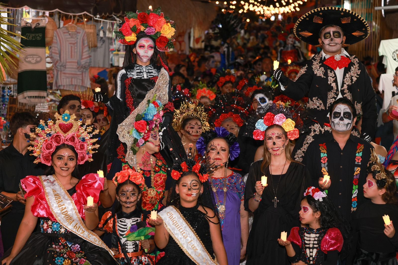 A vibrant celebration of Día de los Muertos featuring participants in traditional costumes and intricate makeup, holding candles and flowers in a lively atmosphere.$#$ CAPTION