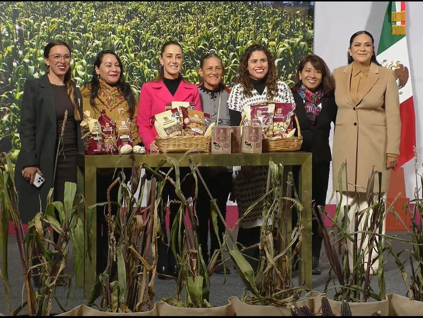 a group of women standing together behind a table filled with agricultural products with corn plants in the background