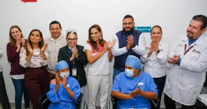 A group of healthcare workers and supporters gathered together, clapping and smiling in a celebratory atmosphere. Two individuals in medical scrubs are seated in front, while others stand behind them. The setting appears to be a healthcare facility.$#" CAPTION
