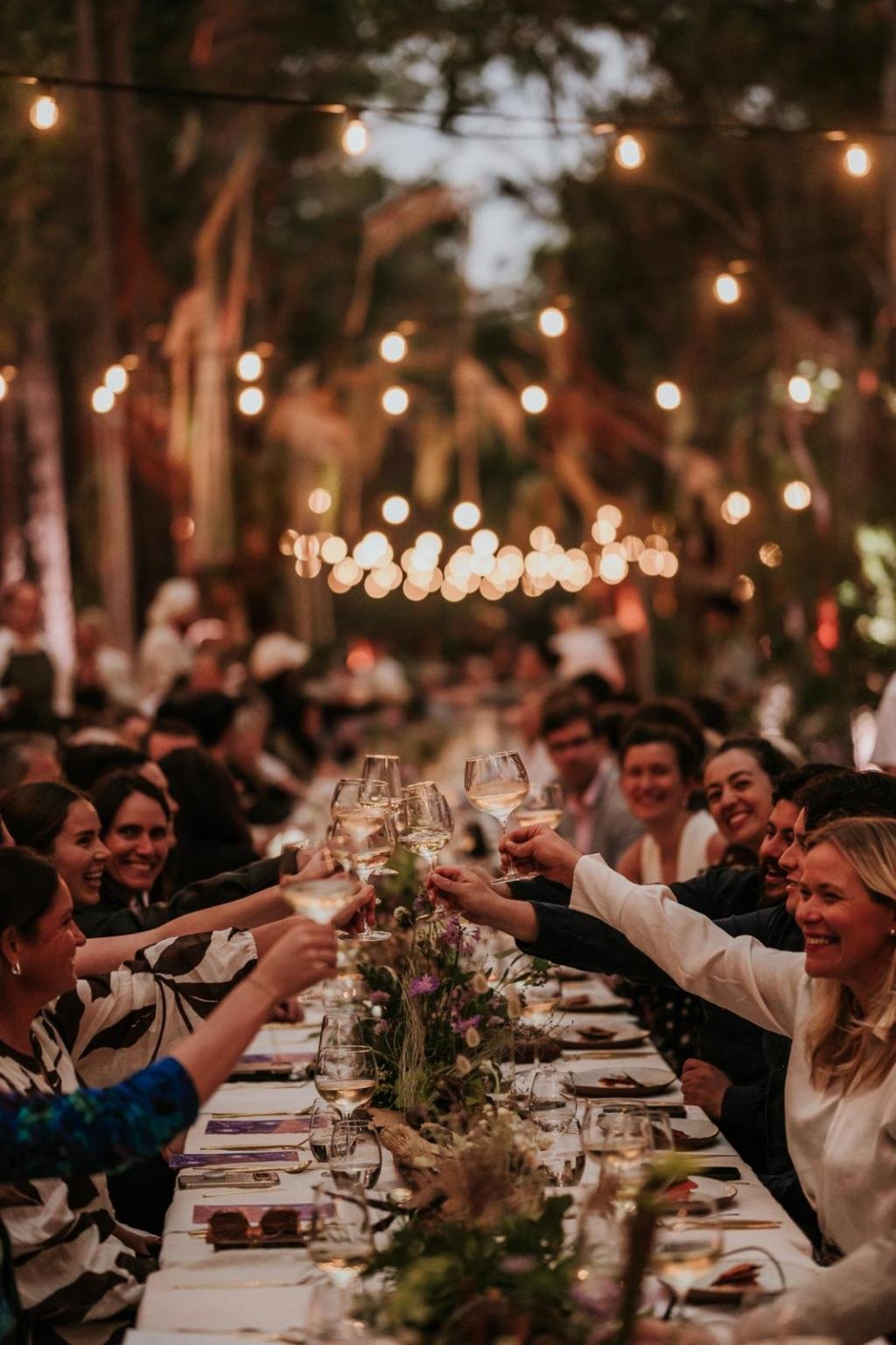 A festive outdoor dinner with people toasting with wine glasses among twinkling lights and floral centerpieces.$# CAPTION