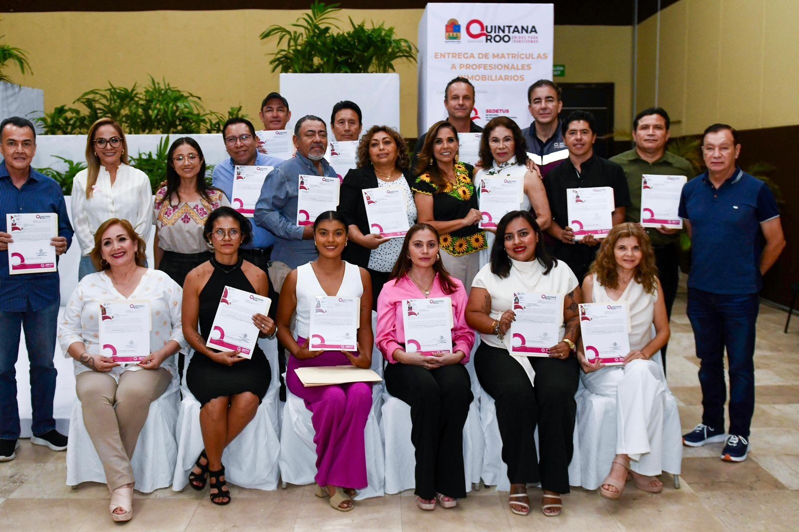 A group of people holding certificates at a certification ceremony for real estate professionals in Quintana Roo, Mexico. The event backdrop displays the logo of Quintana Roo and the name of the institution.