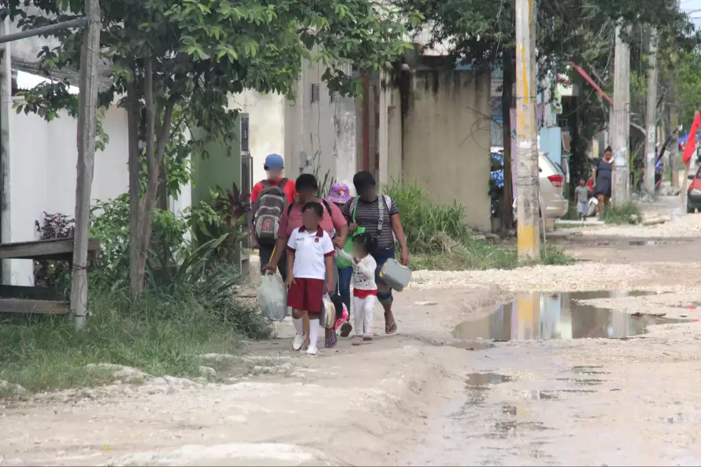 group of children and adults walking down a dirt road in a residential area with greenery and puddles around