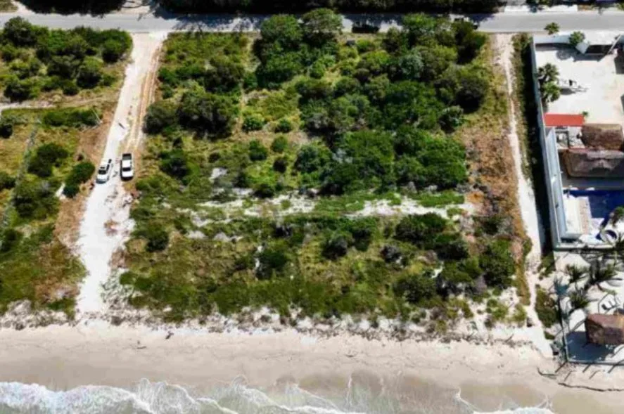 Aerial view of a vacant coastal land area with some vegetation and a nearby beach. Two vehicles are parked on a dirt path leading to the property.$# CAPTION