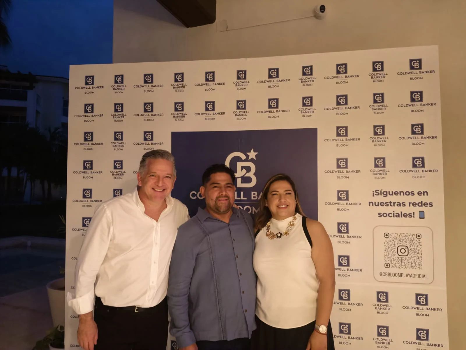 Three individuals posing in front of a branded backdrop at an event for Coldwell Banker Bloom