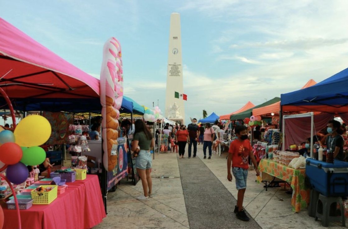 A vibrant outdoor market featuring colorful tents and a tall monument in the background. People are seen walking through the stalls.
