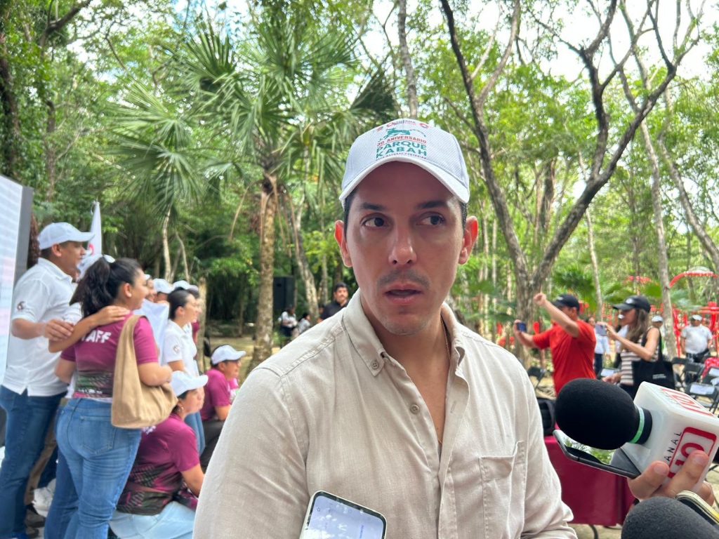 A man in a light shirt and baseball cap speaks at a community event in a park surrounded by trees and people.$#$ CAPTION