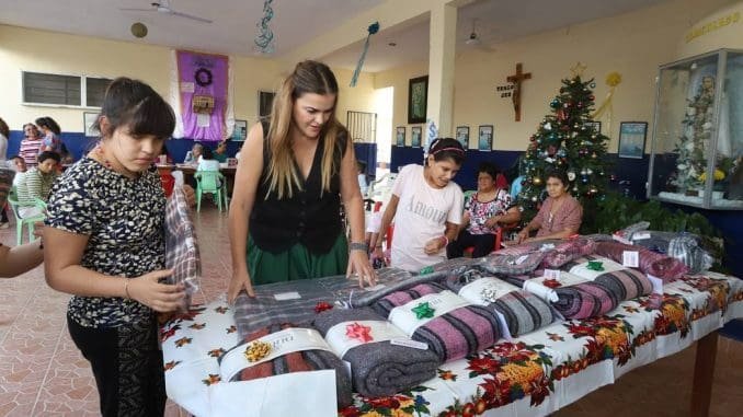 A woman and two girls are arranging wrapped gifts on a table during a community holiday event with decorations in the background