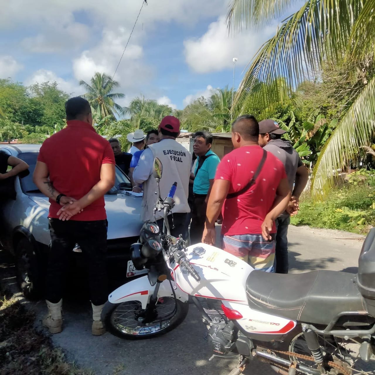 A group of people gathered around a car during a community meeting, with some individuals in discussion and a motorcycle parked nearby.$#$ CAPTION