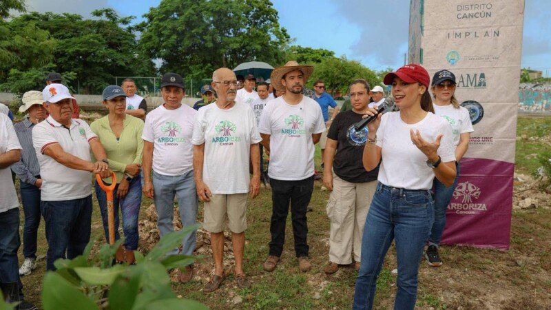A group of people gathered at a tree planting event in Cancun, with a woman speaking to the audience while others listen attentively. Various banners and greenery are in the background.$# CAPTION
