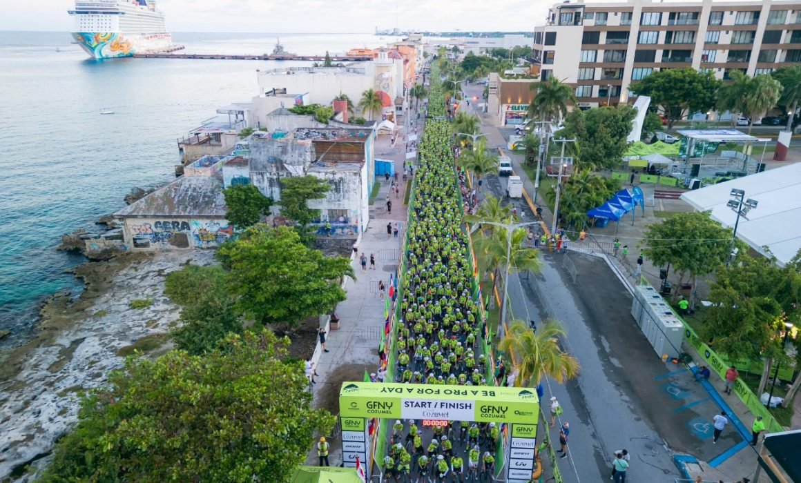 A large group of cyclists in green jerseys gathered at the start/finish line in Cozumel, with buildings and the sea in the background. A cruise ship can be seen in the distance.