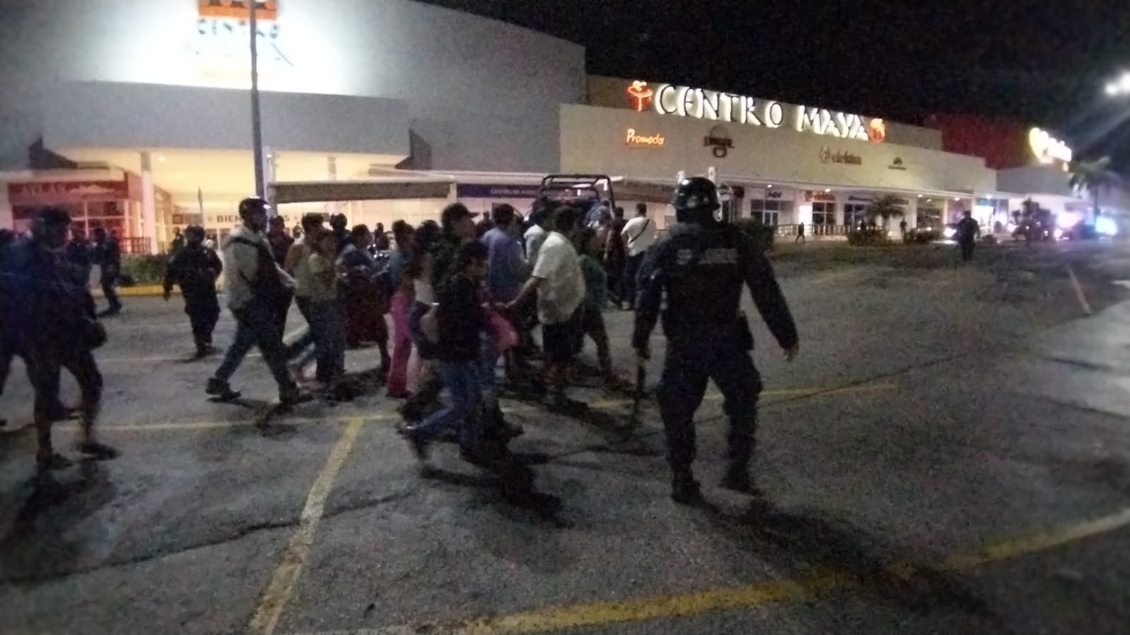 A crowd of people walking outside a shopping center at night, with a police officer in the foreground.