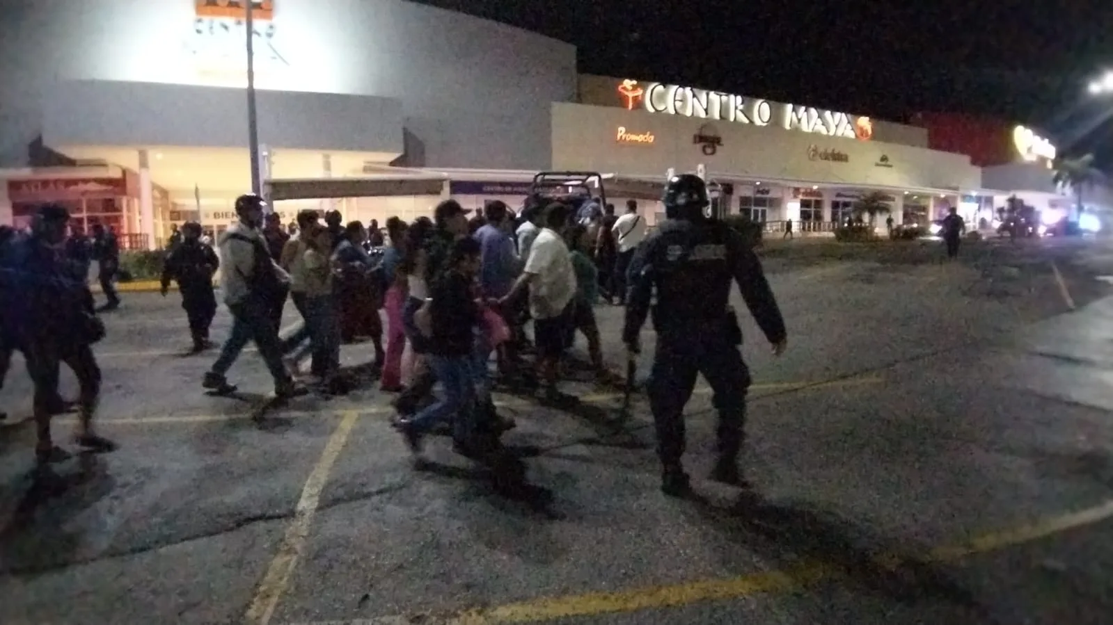 A crowd of people walking outside a shopping center at night, with a police officer in the foreground.