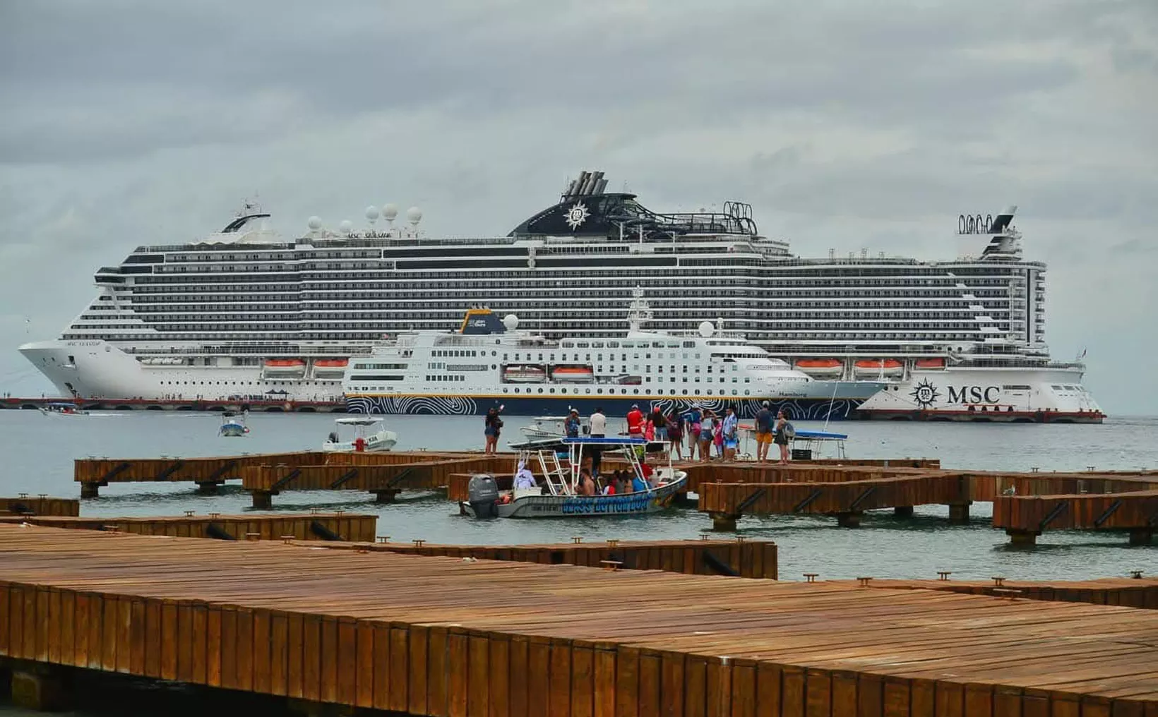 Two large cruise ships docked near wooden piers with people and boats in the foreground.$# CAPTION