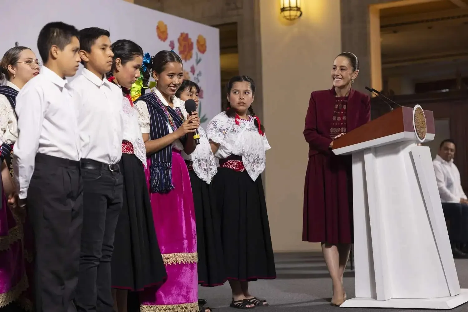 a group of children in traditional attire speaking at a podium during a cultural event with a woman smiling beside them