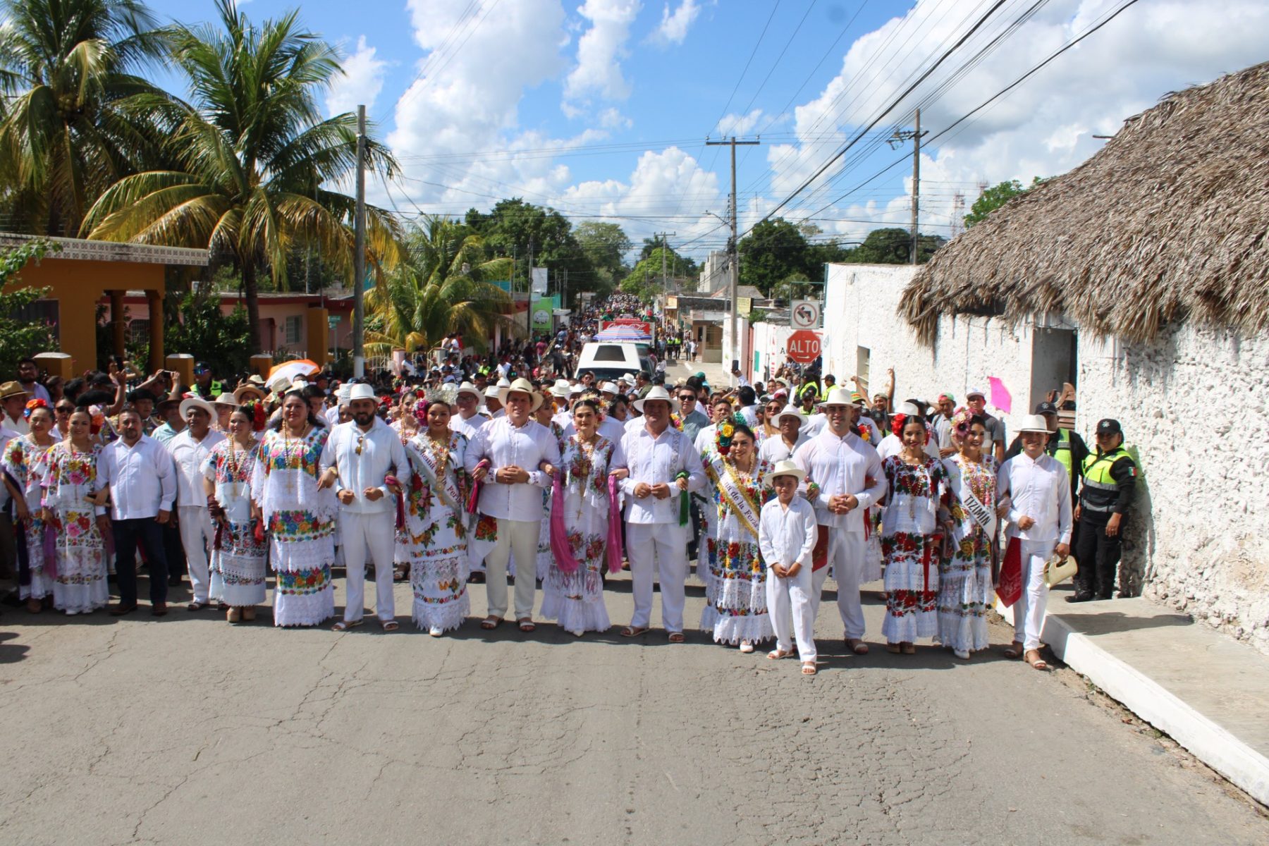 A vibrant group of people dressed in traditional attire during a cultural celebration, with palm trees and a lively crowd in the background.$#$ CAPTION
