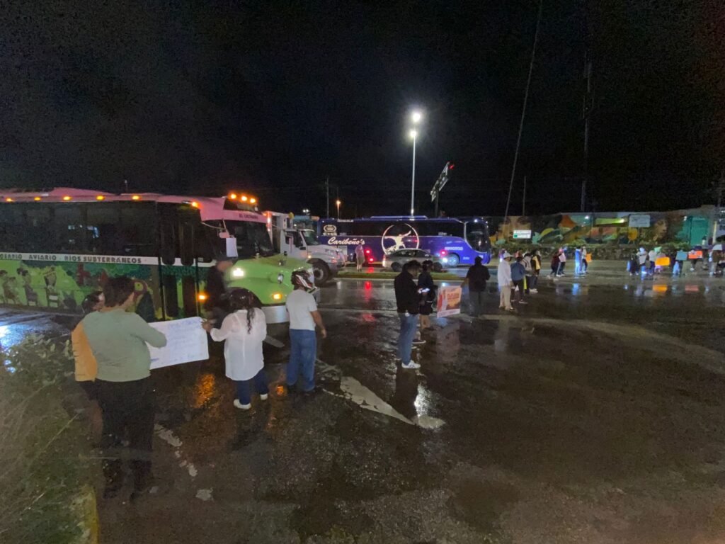 A gathering of people holding signs near parked buses under streetlights at night. The scene is wet, suggesting recent rain.