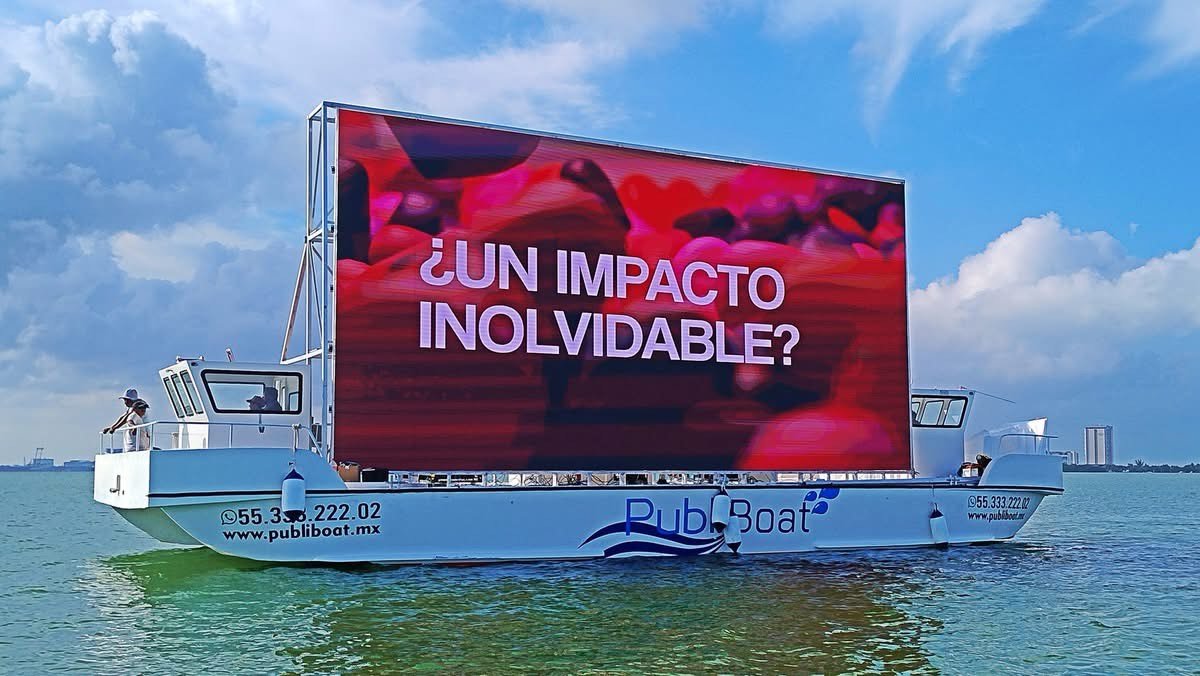 A boat with a large digital screen showing the text "¿UN IMPACTO INOLVIDABLE?" on a body of water, under a partly cloudy sky.$#$ CAPTION