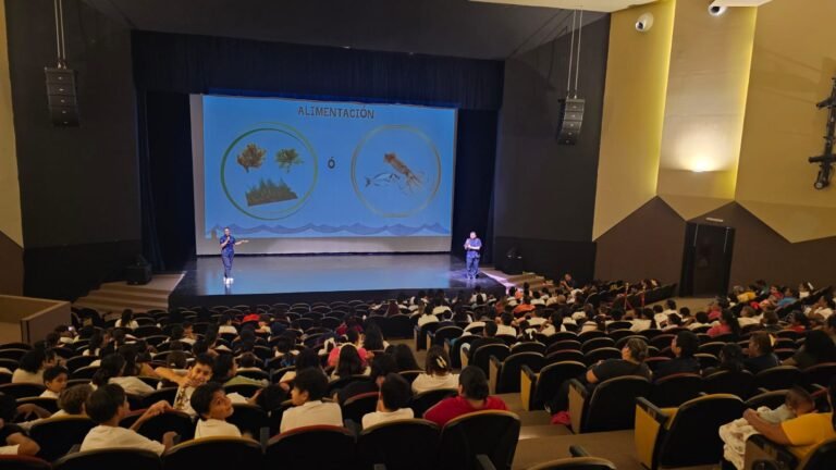 A large audience observes a presentation on nutrition in a theater setting, featuring two images on the screen: one of plants and one of an insect.$# CAPTION