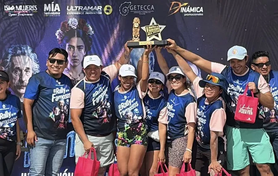 A group of participants celebrating and holding a trophy at the El Rally Famosos event, with a colorful backdrop featuring famous figures. They are dressed in matching shirts and holding pink tote bags.