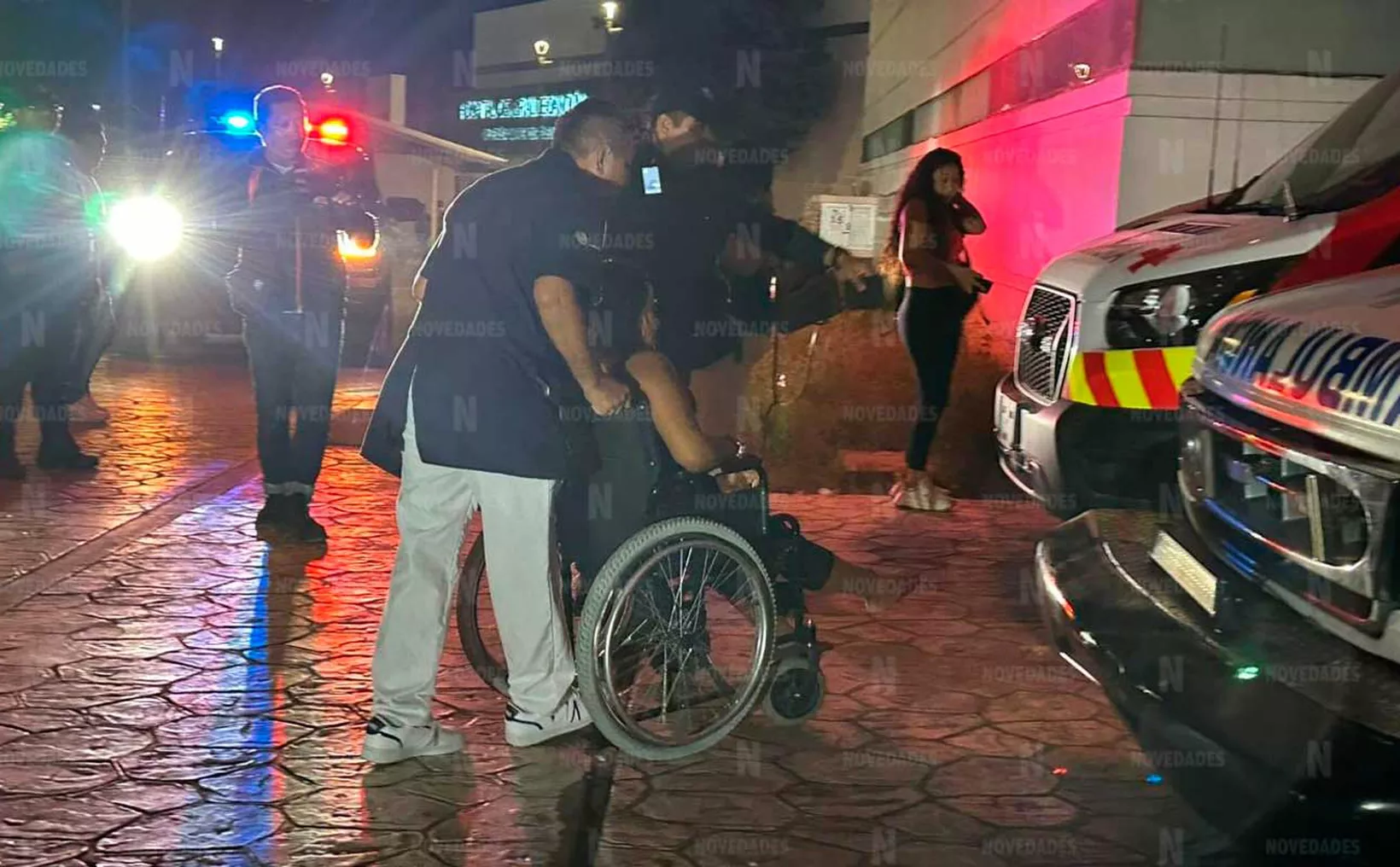 A person in a wheelchair is being assisted by two men near emergency vehicles with flashing lights at night. A woman stands in the background.