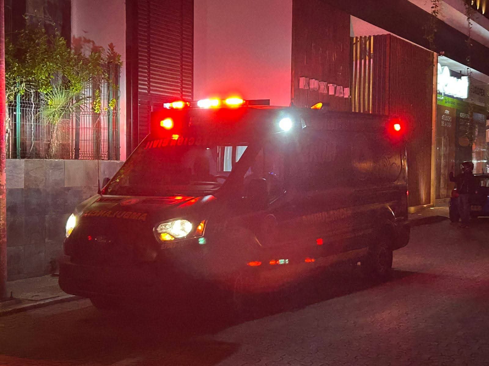 An ambulance with flashing red and blue lights parked on a city street at night, with faint reflections from nearby buildings and trees in the background.$# CAPTION