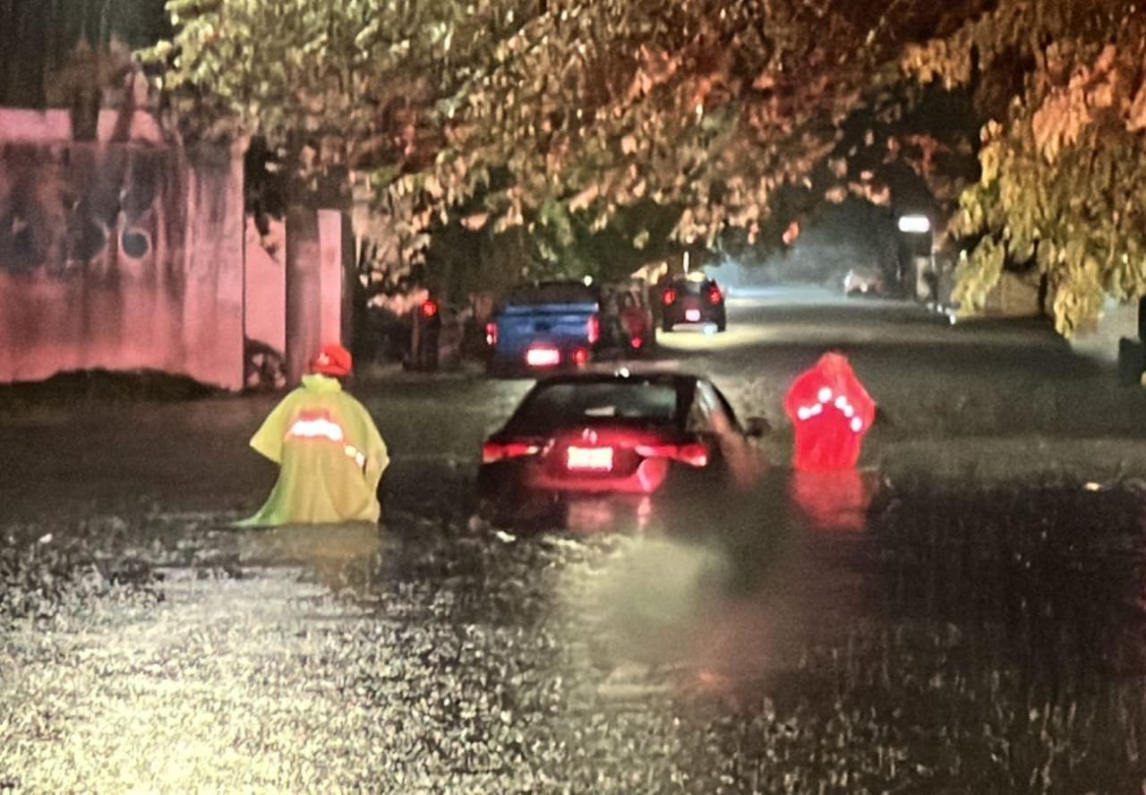 Two individuals in reflective rain gear wading through a flooded street at night, with vehicles partially submerged in the background.$# CAPTION