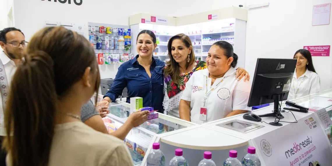 A group of women smiling in a pharmacy, with medications displayed behind the counter. Two women in front are interacting with a customer, while others observe from the back.