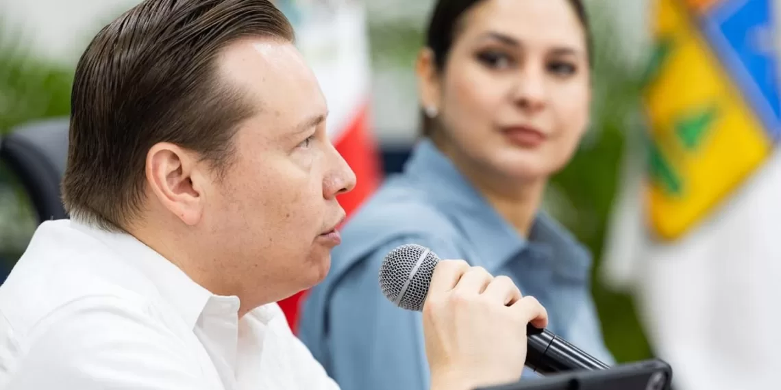 A man speaking into a microphone while a woman listens attentively in the background during a press conference setting.$# CAPTION