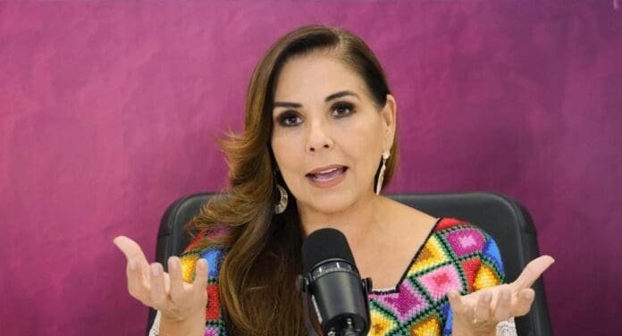 A woman in a colorful embroidered top speaking animatedly at a microphone in front of a vibrant pink backdrop