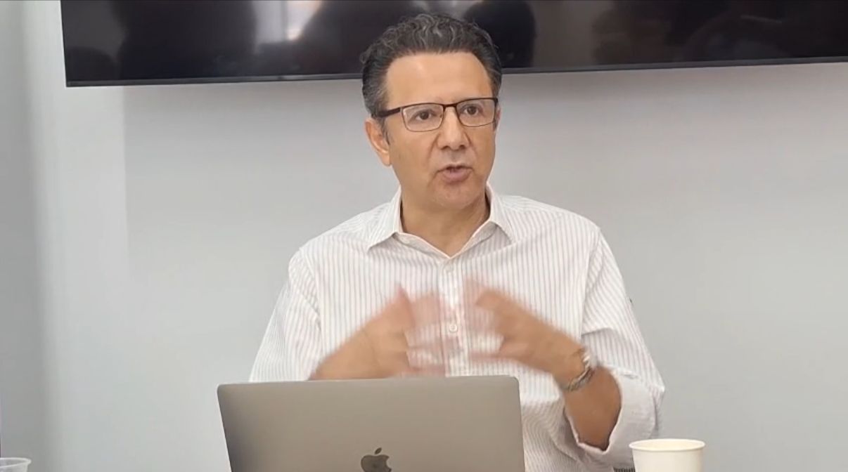 A man with glasses is speaking passionately at a meeting, gesturing with his hands while sitting in front of a laptop and a cup. A television screen is visible in the background.