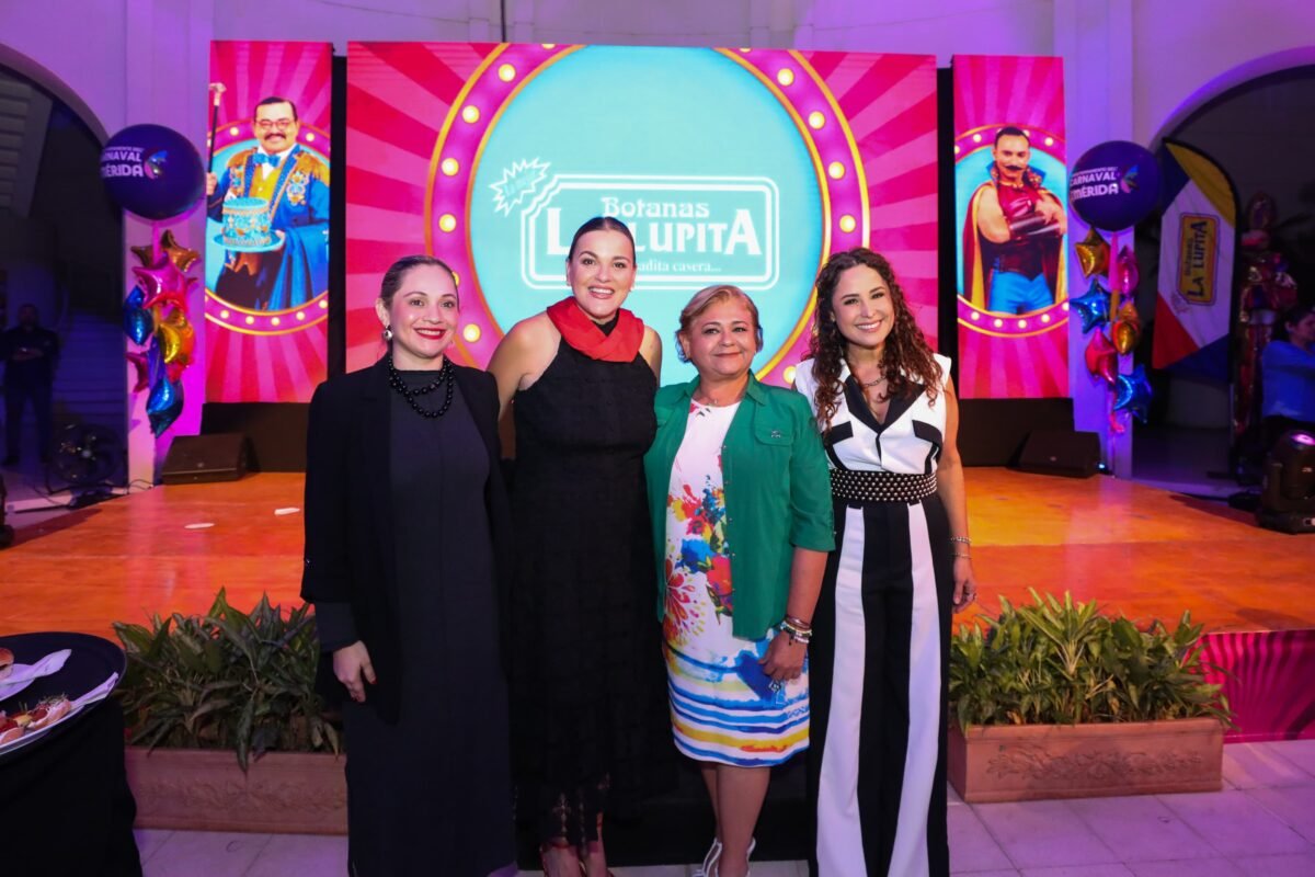 Four women standing together at an event with colorful decorations and a large screen in the background