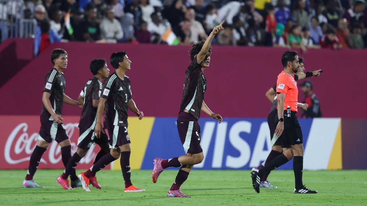 A group of soccer players in brown uniforms celebrates on the field after scoring a goal, while a referee gestures nearby. Spectators can be seen in the background.